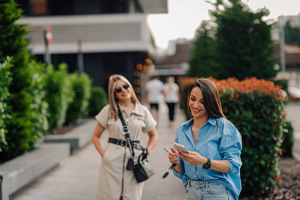 Two smiling businesswomen are walking down the street, one of them using a smartphone, in a modern urban setting