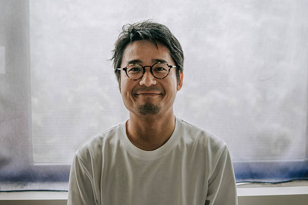 Portrait of a middle-aged Japanese man wearing glasses and a white T-shirt, smiling gently while sitting in front of a softly lit window. The natural backlight creates a bright and calming atmosphere, suggesting themes of mindfulness, inner peace, and everyday simplicity. Ideal for concepts related to wellbeing, authenticity, or home lifestyle.