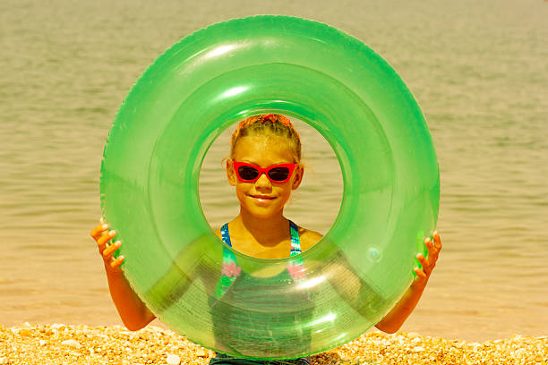 A girl with red braids and red sunglasses playfully looking through a green inflatable ring while standing on a pebble beach, adding a fun and whimsical touch to the seaside scene.