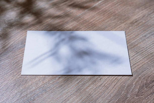 istock Minimalistic white mockup of a blank postcard lying on a wooden surface with soft natural shadows from plants. Template for branding, stationary design, wedding invitations and greeting cards. 2228772497
