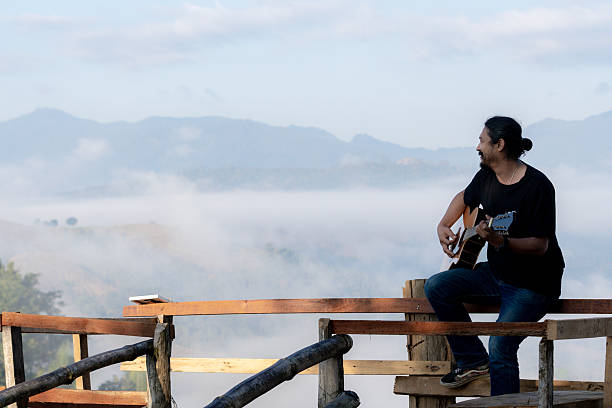 Young man enjoying a peaceful morning, playing guitar on a wooden balcony with warm sunlight and scenic mountain views.