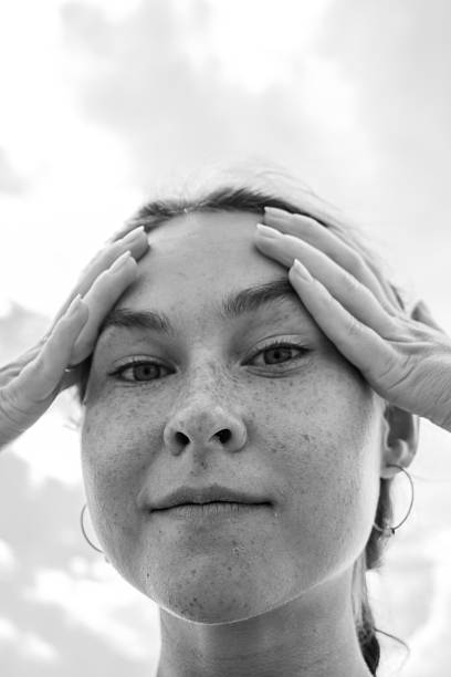 A close-up portrait of a young woman with freckles, gently pressing her forehead with both hands. The background features a cloudy sky, enhancing her contemplative expression.