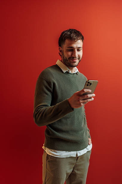 istock Young man using smart phone, smiling and looking at the screen on red background 2228796030