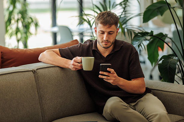 istock Young man using phone and drinking coffee on sofa in modern office 2228796629