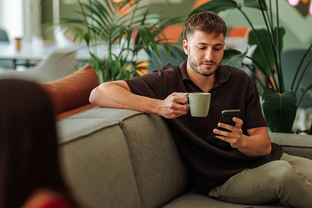 Young Adult Man Sitting on Sofa, Drinking Coffee and Using Smart phone in Modern Office