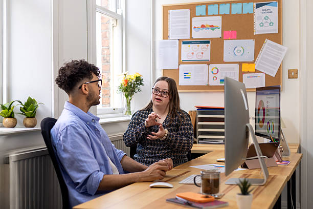 Medium shot of a young adult female with Down syndrome and her mid-adult male colleague sitting at a desk in an office in Newcastle-upon-Tyne, North East England. They are both wearing smart clothing and glasses, discussing work together.Videos similar to this scenario available.