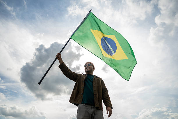 Portrait of cheerful young man looking forward while standing proudly with holding Brasil flag over his head against wind and blue sky, Man holding the Brasil flag celebration