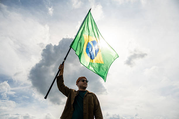 istock Portrait of cheerful young man looking forward while standing proudly with holding Brasil flag over his head against wind and blue sky, Man holding the Brasil flag celebration 2228803659