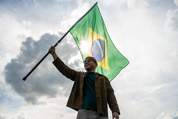 istock Portrait of cheerful young man looking forward while standing proudly with holding Brasil flag over his head against wind and blue sky, Man holding the Brasil flag celebration 2228803924