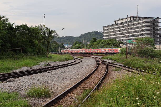 Perak, Malaysia - April 24, 2025 : Curving railway tracks lead through verdant greenery towards a red train stationed near a large building.
