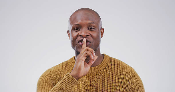 istock Portrait, black man and finger on lips in studio for secret, quiet or privacy gesture for gossip. Happy, confidential and African person with silent hand sign for rumor or mystery by white background 2228811605