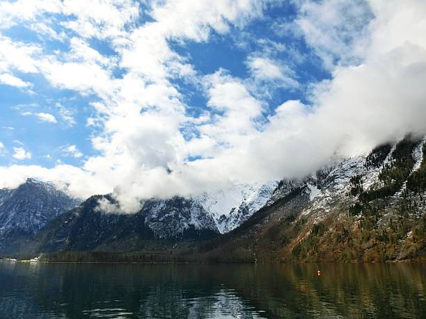 A breathtaking landscape featuring a serene alpine lake surrounded by majestic snow-covered mountains under a partly cloudy sky. The calm water mirrors the rugged peaks and soft clouds, enhancing the scene’s peaceful ambiance. A solitary red buoy adds a subtle focal point and contrast to the natural palette. This image evokes tranquility and awe, ideal for travel, nature, wellness, or environmental themes.