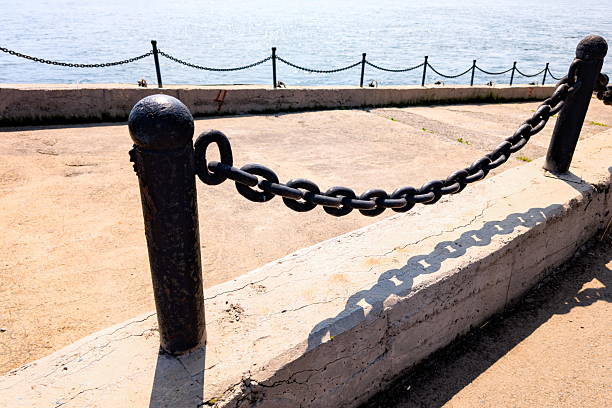 A rusty chain stretches along a concrete barrier by the waterfront, glistening in warm sunlight. The calm sea sparkles behind it, under a bright blue sky with few clouds.