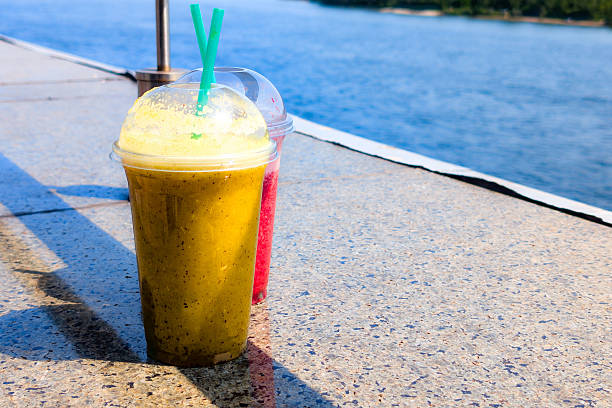 Two colorful smoothies sit on a sunlit surface, showcasing vibrant yellow and red hues. The serene river in the background adds a refreshing touch to the setting.