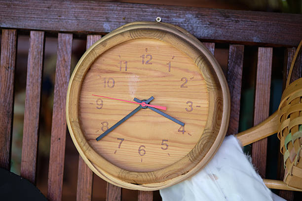 Clock with a wooden frame shows time on a rustic bench in a peaceful outdoor setting. Sunlight highlights the natural grains of the wood and enhances the tranquil atmosphere. Copy space.