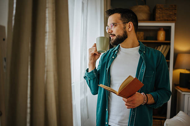 istock Young man drinking coffee and reading a book by the window at home 2228847000