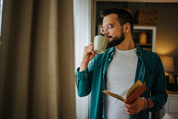 istock Young man drinking coffee and reading a book by the window 2228847003