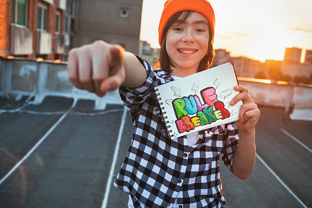 A smiling boy points directly at the camera while holding a “rule breaker” sketch, city skyline glowing behind at sunset.