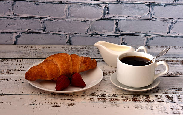 French light breakfast, fresh croissant with strawberries and a cup of black coffee with cream on a wooden table.