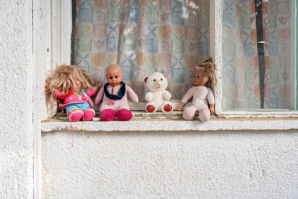 A collection of vintage dolls and a teddy bear are arranged on a windowsill. The toys showcase unique styles, adding charm to the cozy home ambiance. The background features patterned curtains.