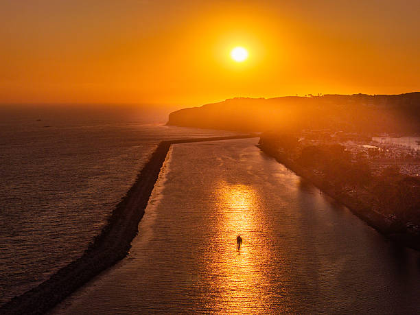 A solitary stand-up paddleboarder glides through the golden water of Dana Point Harbor at sunset. The aerial view captures the peaceful scene and dramatic light reflecting off the Pacific Ocean in Southern California.
