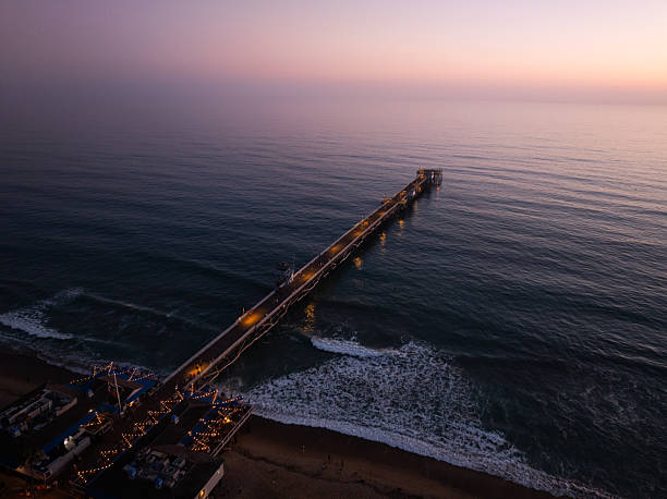 Drone view of the San Clemente Pier extending into the Pacific Ocean during twilight, with warm lights illuminating the structure and gentle waves rolling onto the beach beneath a pastel sky.
