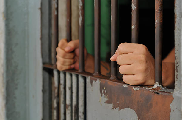 istock prisoner's hands behind bars with black background 2228870219