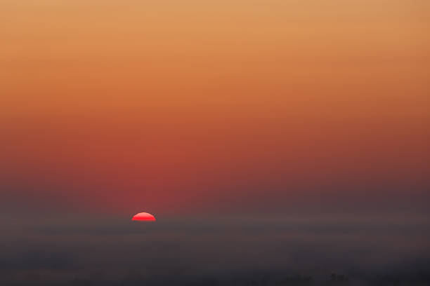 Minimalist sunset with a vivid red sun dipping below a fog-covered horizon, casting a warm gradient across the sky.