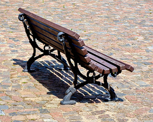 A solitary wooden bench with decorative black metal legs stands on a cobblestone pavement. The sunlight casts a clear shadow of the bench on the stones, emphasizing the textures of the wood and the ground.