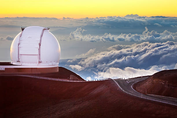 A glowing sunset above the clouds as seen from Mauna Kea, featuring a large observatory dome overlooking the valley below.
