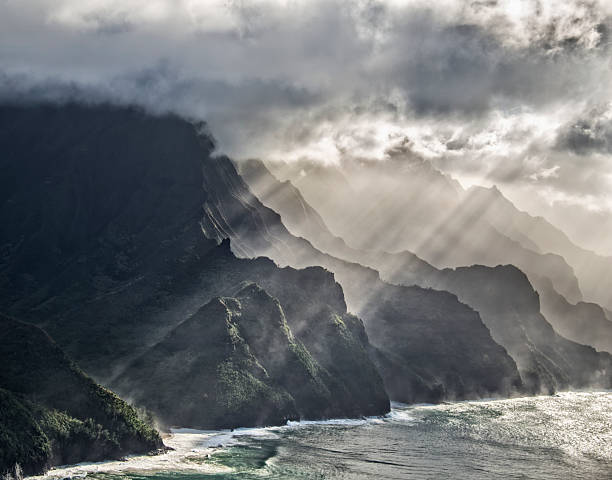 A vivid aerial photo of the Na Pali Coast on Kauai, Hawaii, with sharp cliffs, ocean waves, and streaming sunlight.