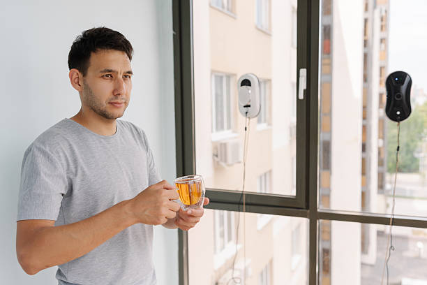 Smart home technology enhancing daily living, window-cleaning robot operating quietly while resident male relaxing with morning tea in contemporary apartment interior. Convenient electronic device
