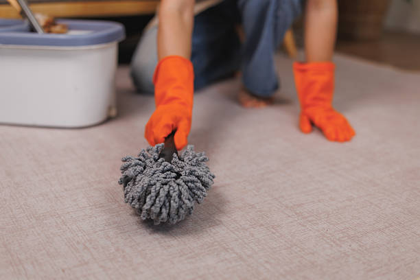 Stock photo showing close-up view of an unrecognisable person wearing casual clothing of a blue and white striped polo shirt, feather patterned shorts and a pair of thong sandals to clean and tidy an empty residential property by sweeping up dust and dirt, into a heap on the tiled flooring, with a short-handled broom.