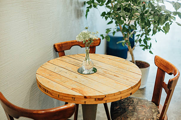 A round rustic wooden table with two vintage chairs features a glass vase of white wildflowers, set beside a potted indoor plant in a cozy, natural-lit interior