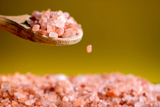 Extreme close up studio shot of a spoon filled with a himalayan salt, being poured. Close up shot of falling crystals of Himalayan salt into a wooden bowl, with yellow background. Studio shot. No visible human presence.