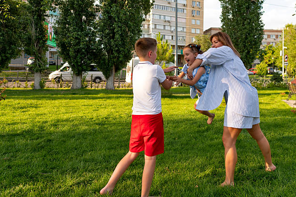Mother swings her squealing daughter toward her waiting brother on a bright grassy lawn, all three laughing and moving fast in a playful summer park scene.