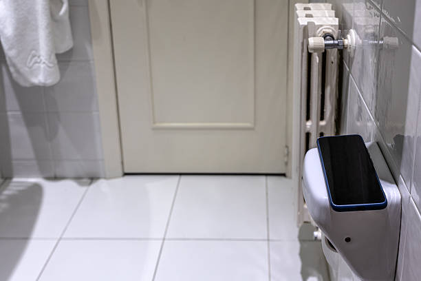 Spain. Contemporary bathroom with a mobile phone resting on the ceramic toilet paper dispenser. The bathroom is complemented by a white door, a radiator, and a minimalist tiled floor, creating a clean aesthetic.
