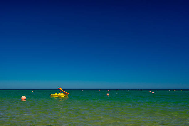 Relaxed summer holiday atmosphere with crystal clear sea waters and a yellow leisure boat with a red slide floating in the calm waters of Barril beach - Santa Luzia - Tavira - Algarve - Portugal