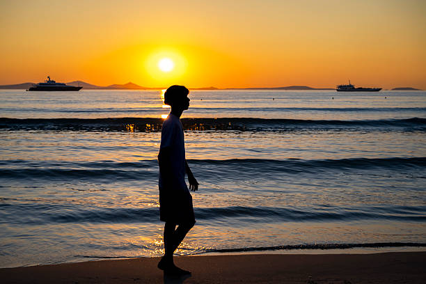 Silhouette of a person walking by the Aegean Sea at dusk in Naxos, Greece. Warm tones and calm waters create a serene coastal moment.