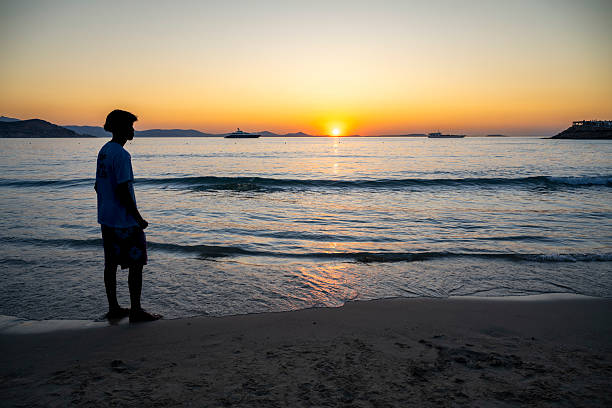 A man in profile walks along the shoreline as the sun sets behind distant hills and anchored yachts near Naxos. Peaceful and contemplative.