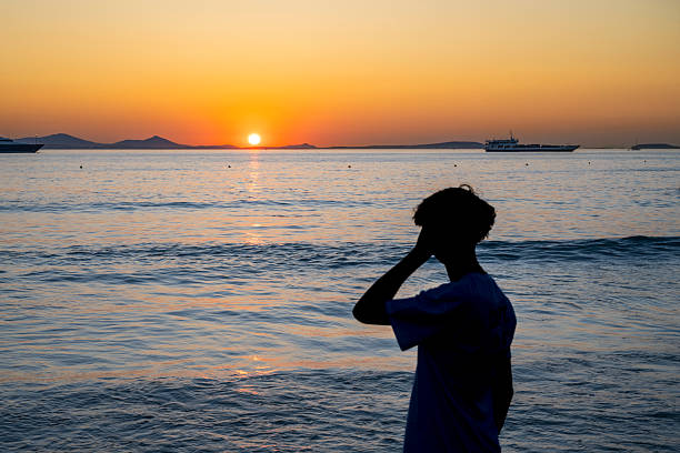 Silhouette of a person watches the sun set over calm waters in Naxos, Greece, as yachts cruise in the distance. A serene depiction of wealth, solitude, and lifestyle.