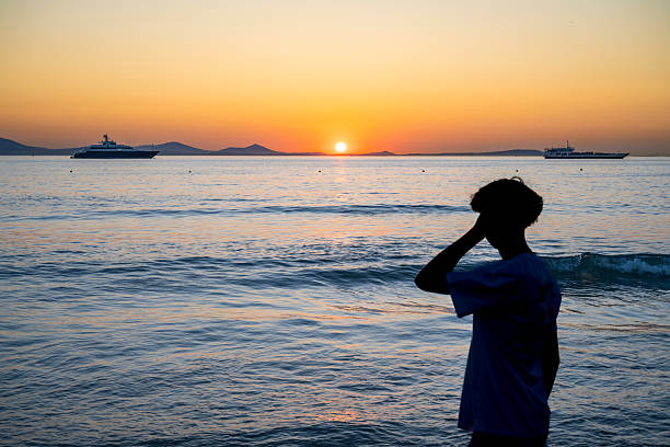Luxury yachts glide across the sea during a vivid Greek sunset, with an unrecognizable figure silhouetted in thought. A lifestyle scene blending nature, leisure, and exclusivity.