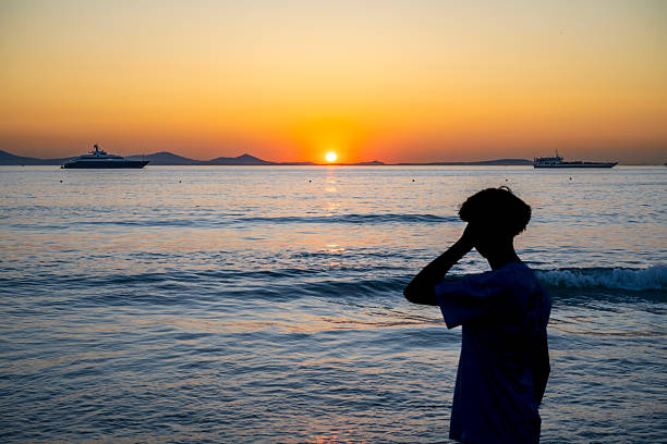 A lone figure stands on the beach at sunset, facing luxury vessels anchored offshore. A tranquil, anonymous moment evoking wealth and introspection.