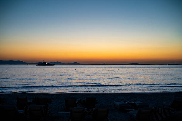 An empty beach faces a calm sea as twilight falls over Naxos. A yacht glows in the distance beneath a fading orange sky.