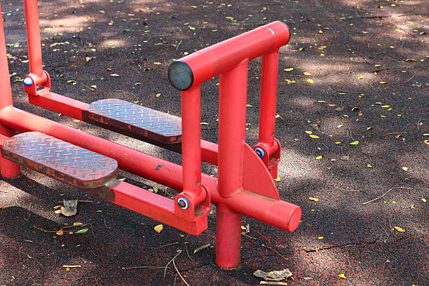 A close-up of a red metal air walker machine at an outdoor gym. the equipment sits on a dark rubber floor covered in leaves.