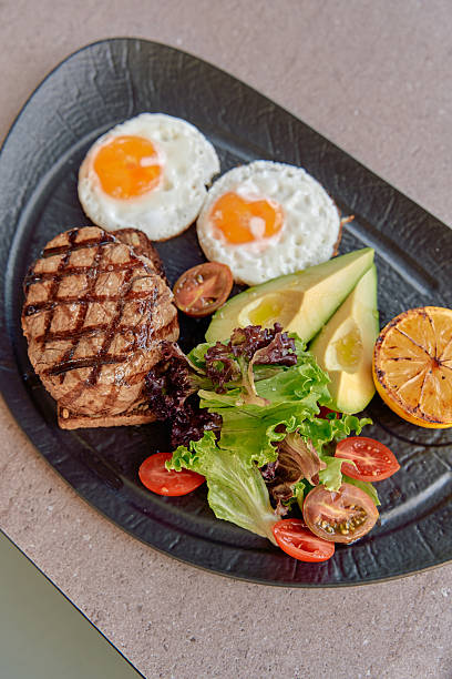 A Deliciously Grilled Steak Platter Accompanied by Eggs, Fresh Greens, Avocado, and a Zesty Lemon Slice on a Black Plate