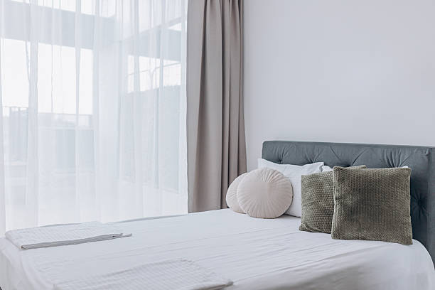 istock Bright bedroom with large window, beige curtains, and round decorative pillows on white bed. 2228933098