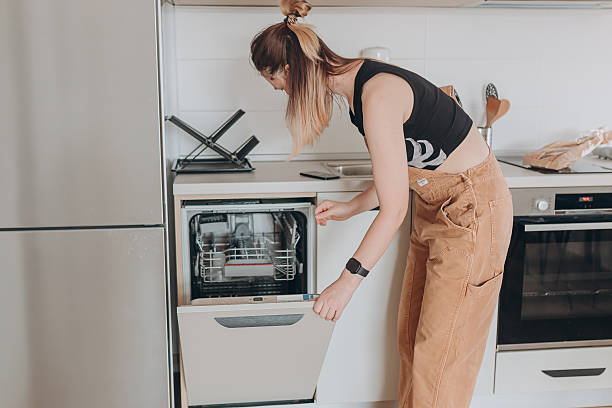 istock Woman closing dishwasher after loading clean white dishes in modern home kitchen with neutral tones. 2228933105