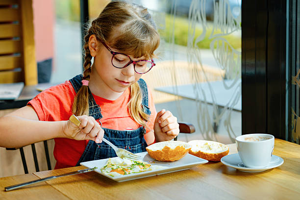 Adorable little elementary school girl with glasses having breakfast in a cozy cafe. Child eating fried eggs, buttered bread and drinking hot cocoa. Real life, real people moment.