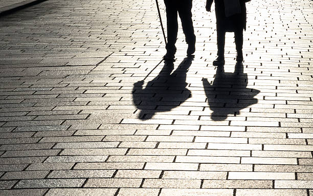 Stark shadows cast during November as an elderly man walks with a walking stick with his partner.